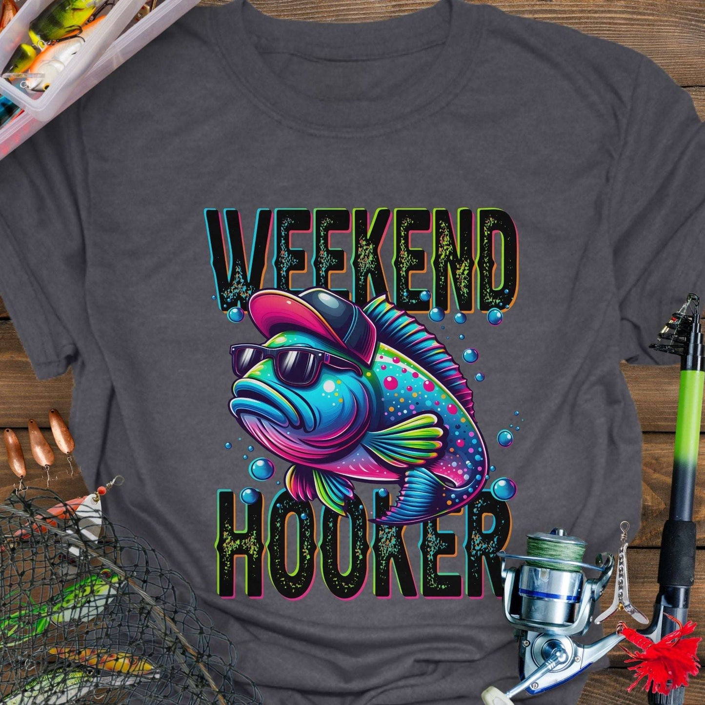 Gray t-shirt with a colorful fish graphic and 'Weekend Hooker' text on a wooden surface with fishing equipment.