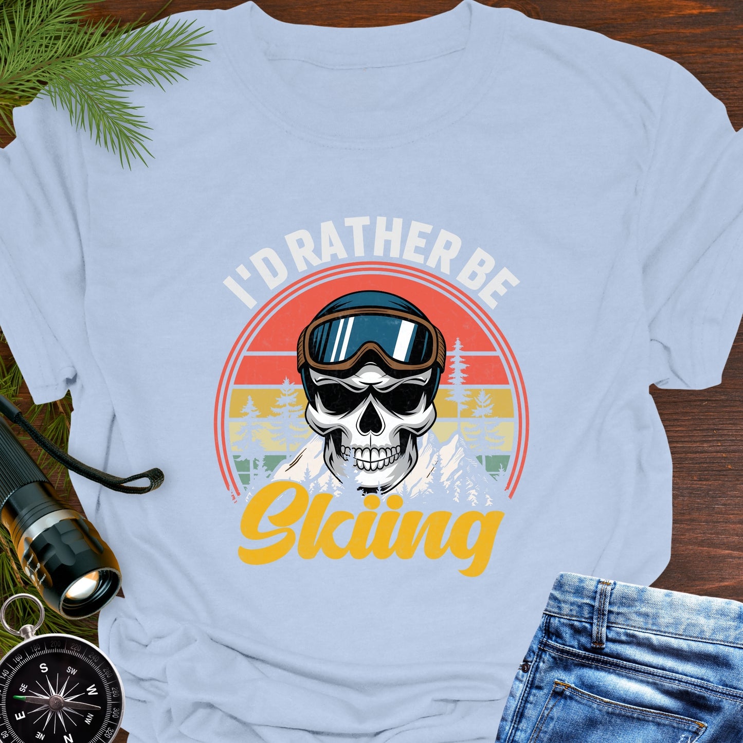 Light blue T-shirt graphic showing a skeleton head wearing ski goggles in front of a retro sunset with mountains and trees, with text that reads “I’d Rather Be Skiing.