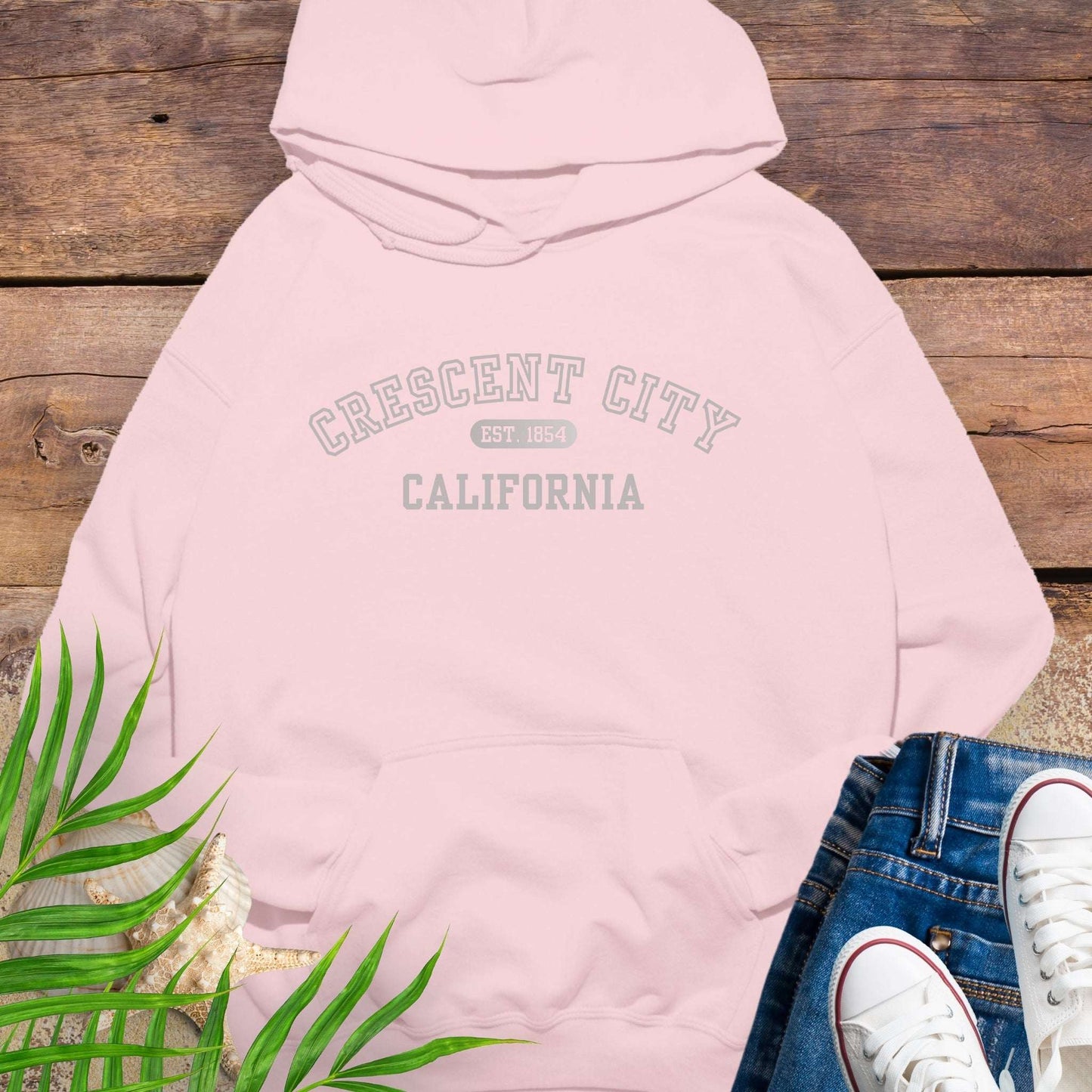 Pink hoodie with 'Crescent City, California Est. 1854' text on a wooden surface with greenery and white sneakers.