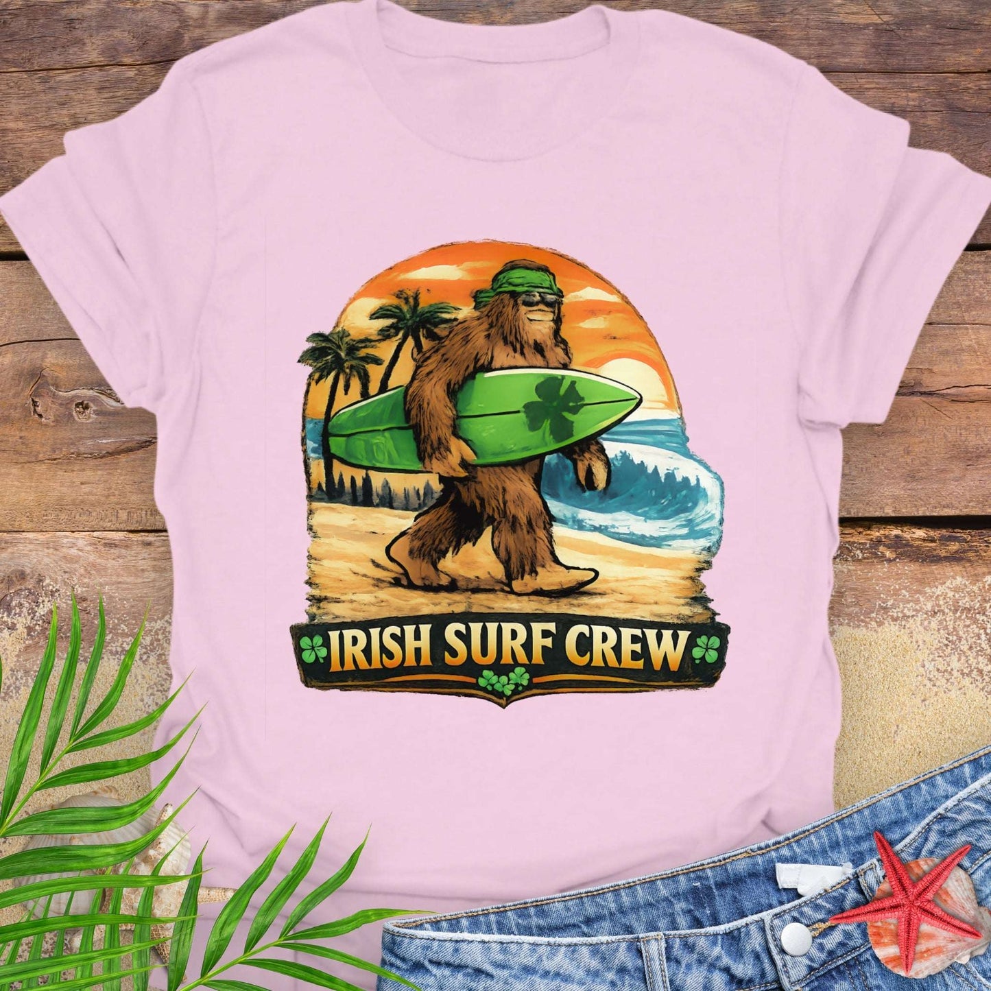Pink t-shirt with Bigfoot carrying a shamrock-decorated surfboard at the beach, with text “Irish Surf Crew”