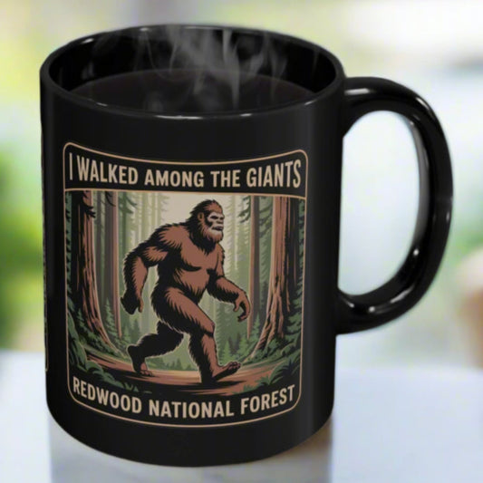 Black mug with a Sasquatch design and text 'I Walked Among the Giants, Redwood National Forest' on a blurred background.