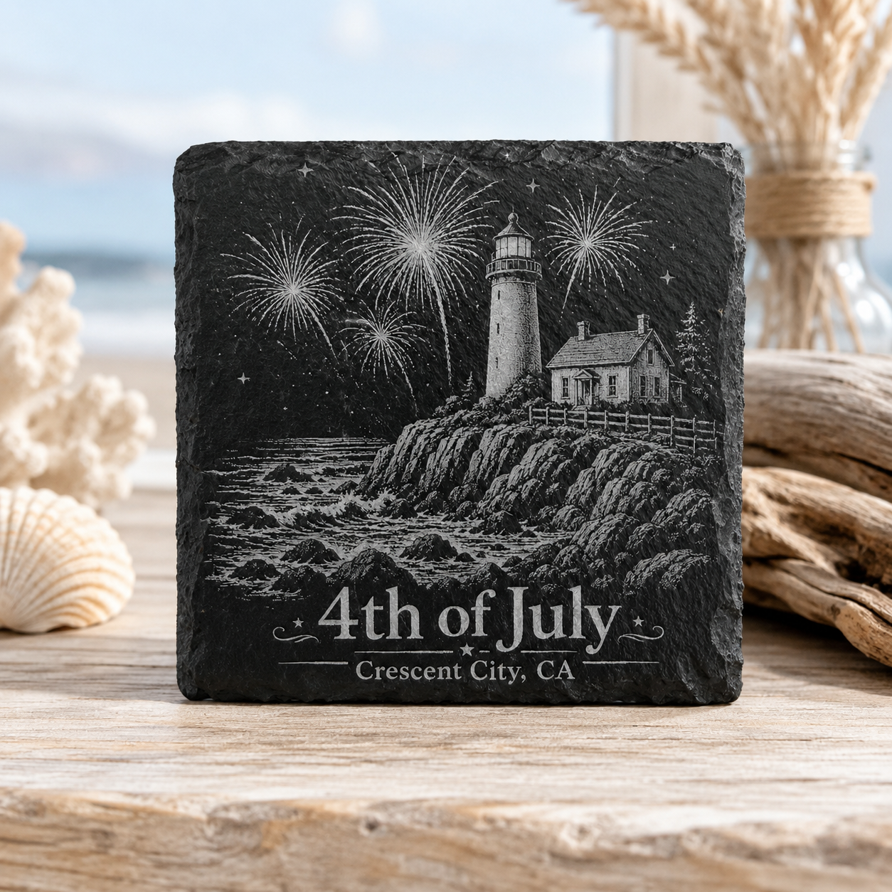 A square black slate coaster featuring a white engraved illustration of a lighthouse, fireworks, a house, and the text '4th of July' and 'Crescent City, CA', displayed on a wooden surface with beach-themed decor in the background.