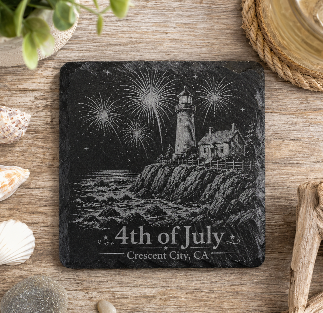 A square black slate coaster featuring an engraved lighthouse, fireworks, rocky shoreline, and the text '4th of July' and 'Crescent City, CA', displayed on a rustic wooden surface with shells and decorative props around it.