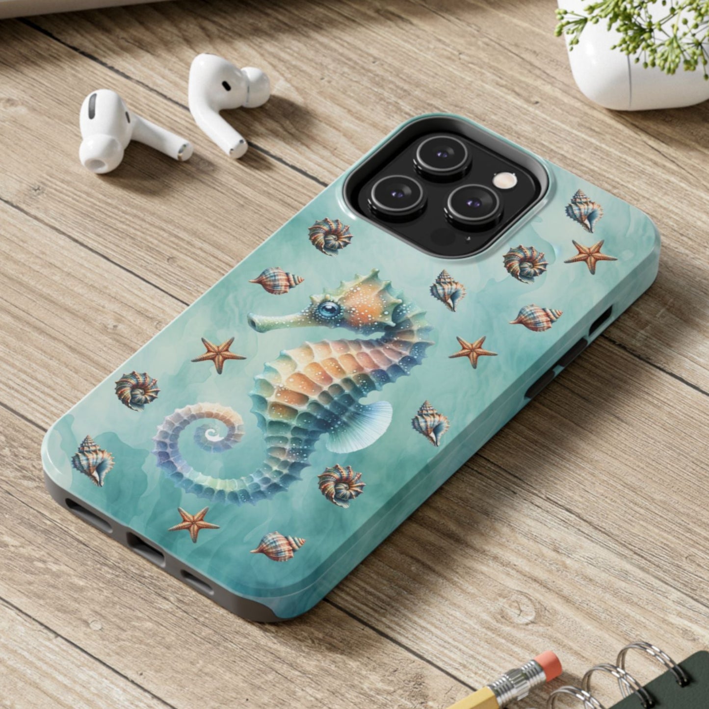 Phone case with seahorse design on a wooden surface with laptop, earbuds, and notebook.