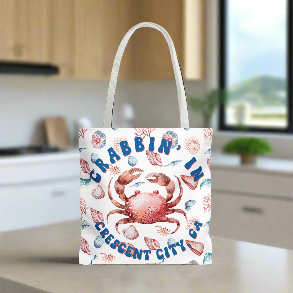 White Tote bag featuring a crab surrounded by seashells and underwater life in blue and soft red tones with the phrase "Crabbin' in Crescent City, CA.
