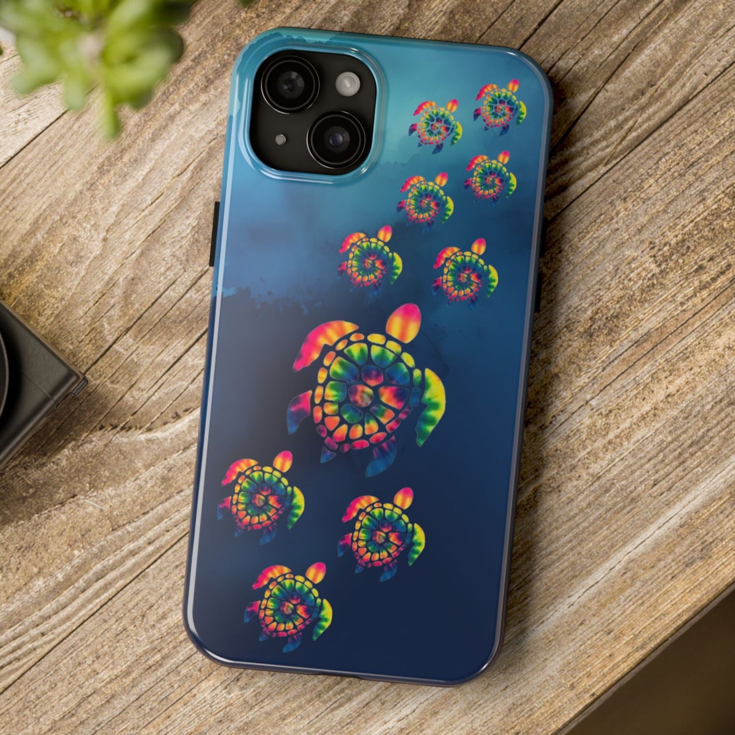 Phone case with tie-dye pattern on a wooden surface next to a camera and plant.