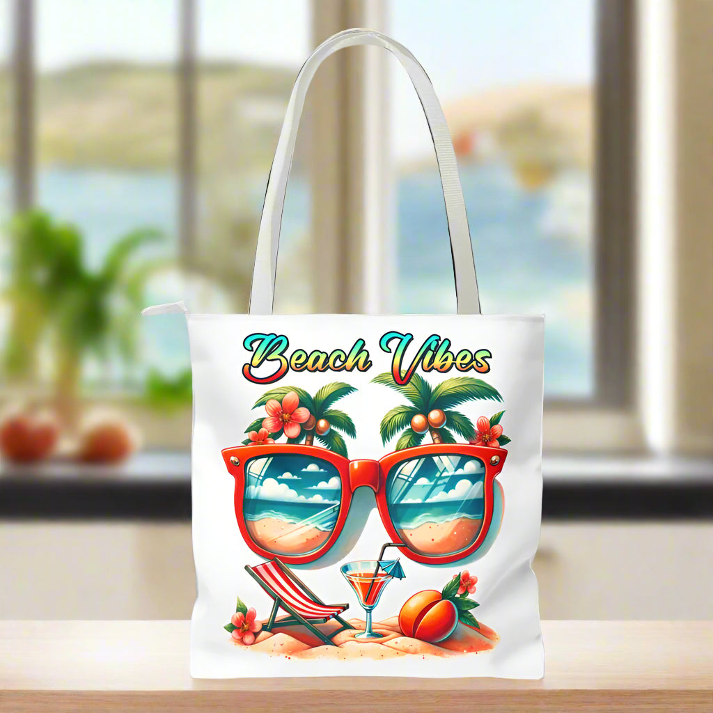 White Tote bag featuring sunglasses with ocean and beach scene reflections, palm trees coming out of the glasses, a beach chair and cocktail on the sand, with the phrase "Beach Vibes" in a retro rainbow gradient font.