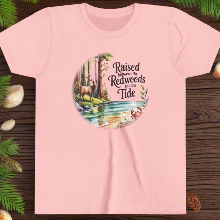 Pink t-shirt with nature-themed graphic with elk among redwoods and stream with a crab and text Raised between the Redwoods and the Tide.