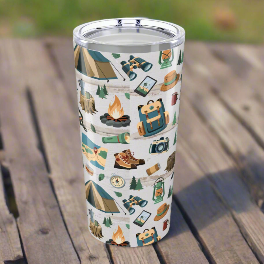 Back of 20oz tumbler with light wood background and repeating pattern of hiking boots, tents, campfires, flashlights, maps, and backpacks; glossy finish with secure lid.