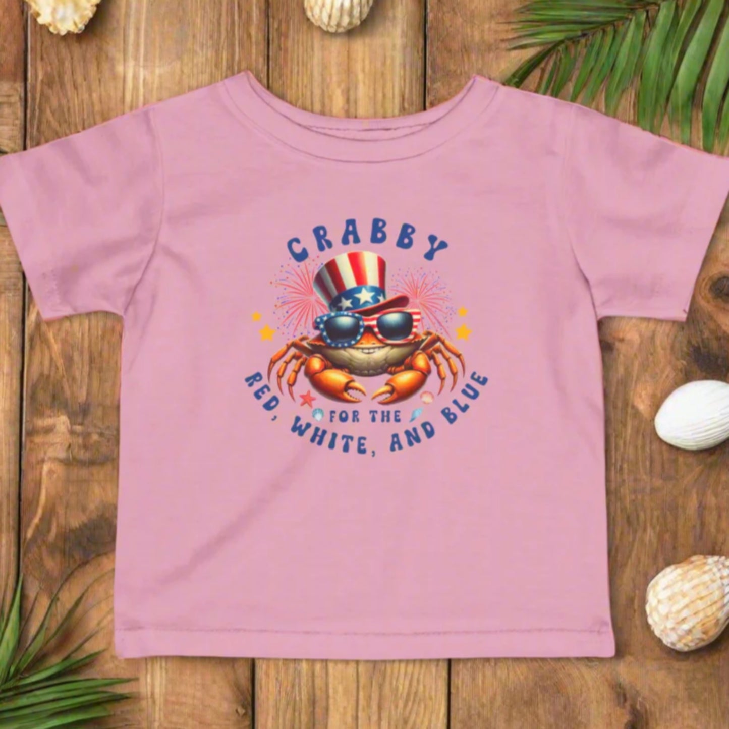 PInk Infant t-shirt with a crab wearing an American flag top hat surrounded by fireworks and seashells with the phrase “Crabby for the Red, White, and Blue.”