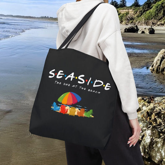 Black 18" x 18" tote bag with the word “Seaside” in rainbow-style letters with colored dots between each letter, underneath says The One at the Beach with five rainbow Adirondack chairs, and a beach umbrella on sand graphic.