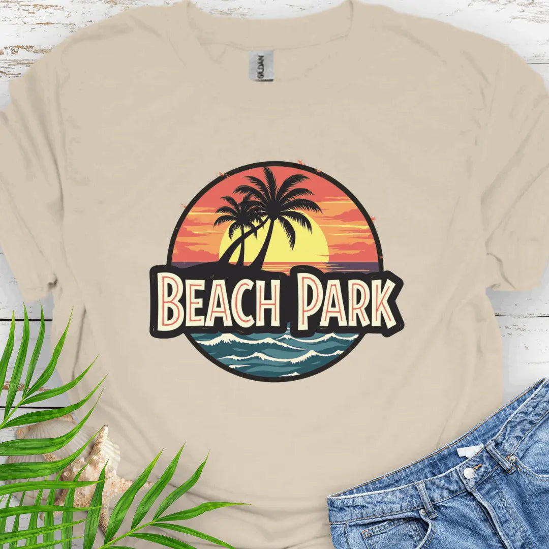 Beige T-shirt with a vintage-style logo inspired by Jurassic Park, but with a beach theme featuring a circular emblem with a bold, retro font spelling 'BEACH PARK' in the center. The dinosaur silhouette is replaced with a tropical scene of palm trees.