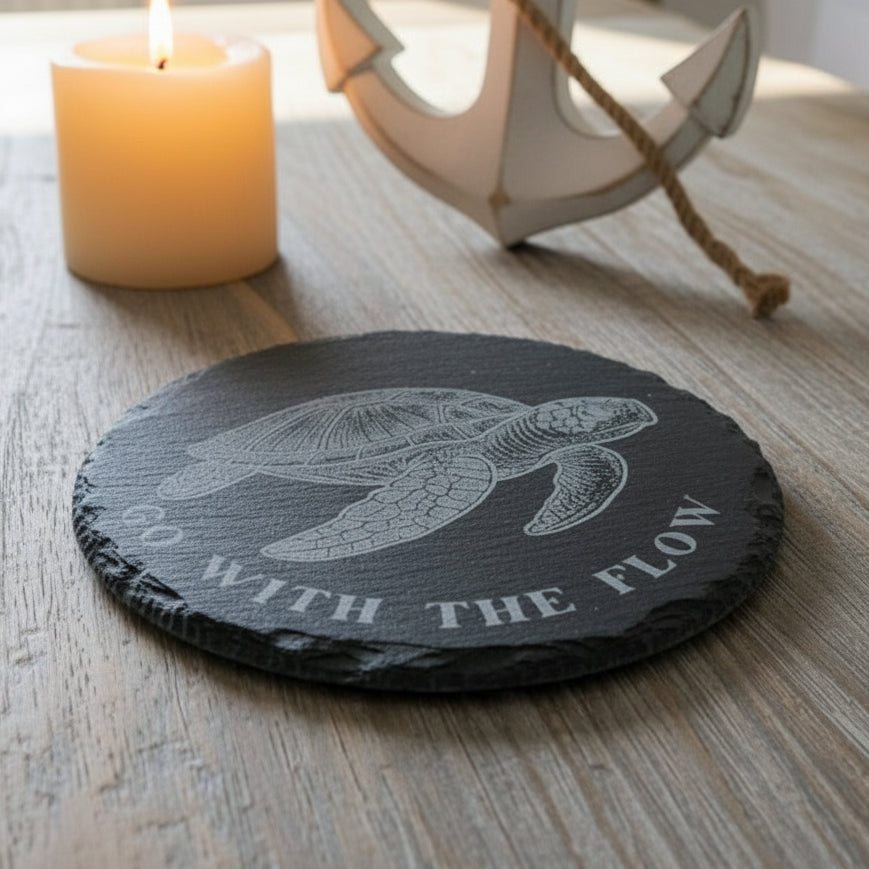 Slate coaster with turtle design and 'Go with the Flow' text on a wooden surface with a candle and anchor in the background.