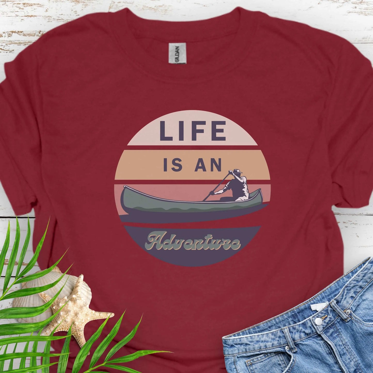 Cardinal Red t-shirt featuring a retro sunset design with a man paddling a canoe and the phrase "Life is an Adventure" in bold, vintage-style font. Perfect for outdoor lovers, kayakers, and adventure seekers.