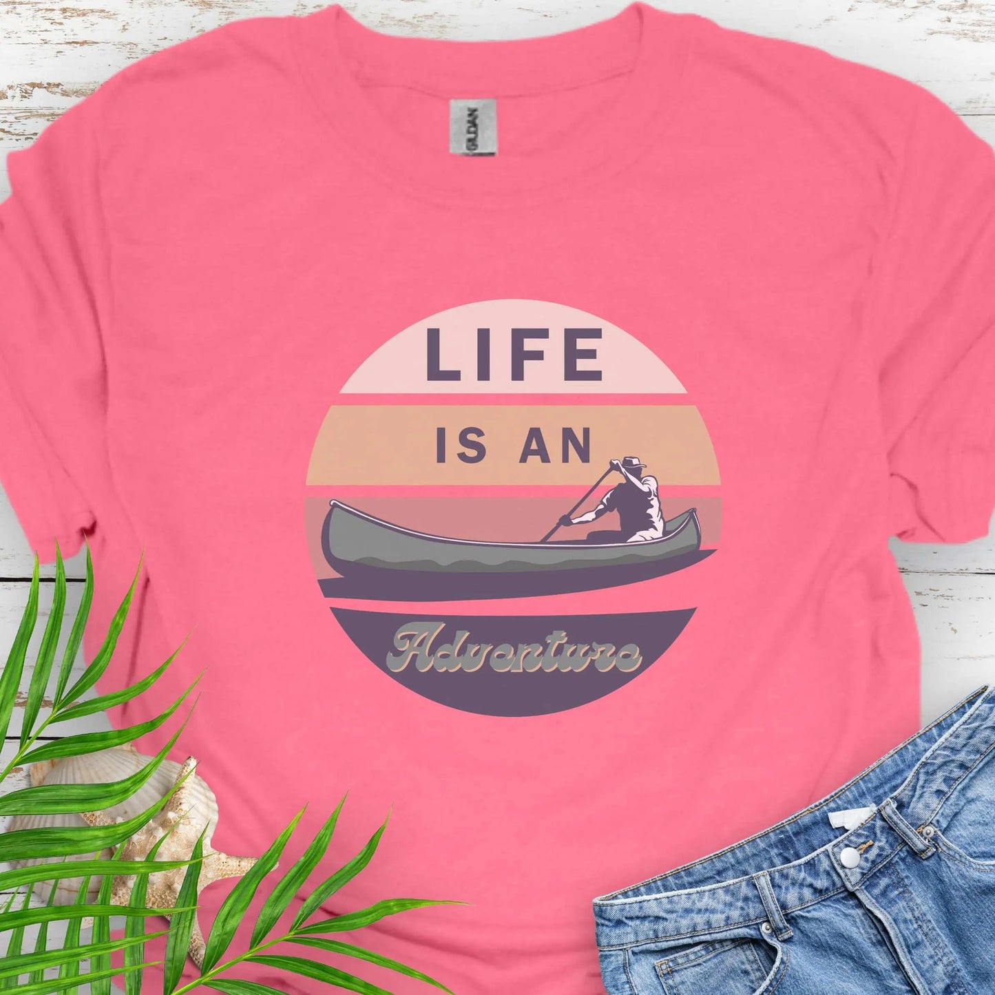 Coral Silk t-shirt featuring a retro sunset design with a man paddling a canoe and the phrase "Life is an Adventure" in bold, vintage-style font. Perfect for outdoor lovers, kayakers, and adventure seekers.