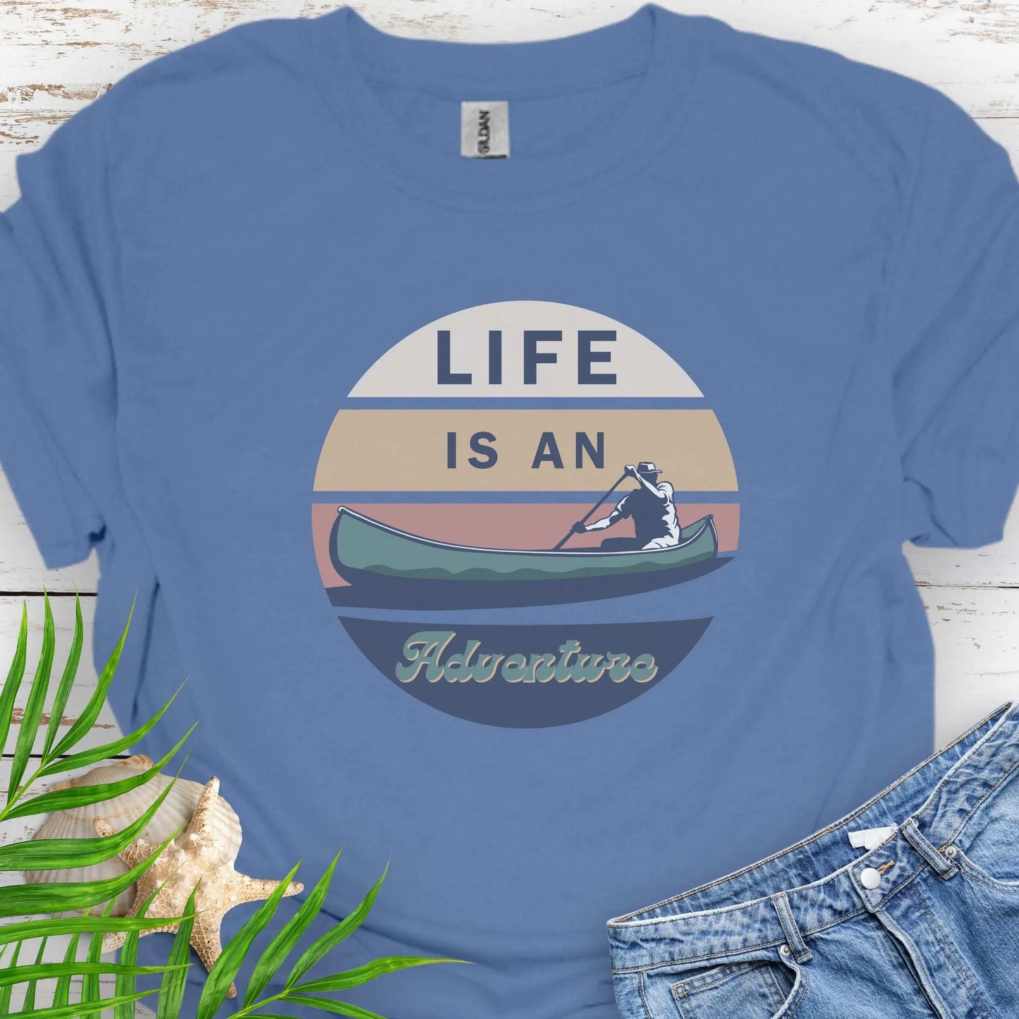 Indigo Blue t-shirt featuring a retro sunset design with a man paddling a canoe and the phrase "Life is an Adventure" in bold, vintage-style font. Perfect for outdoor lovers, kayakers, and adventure seekers.