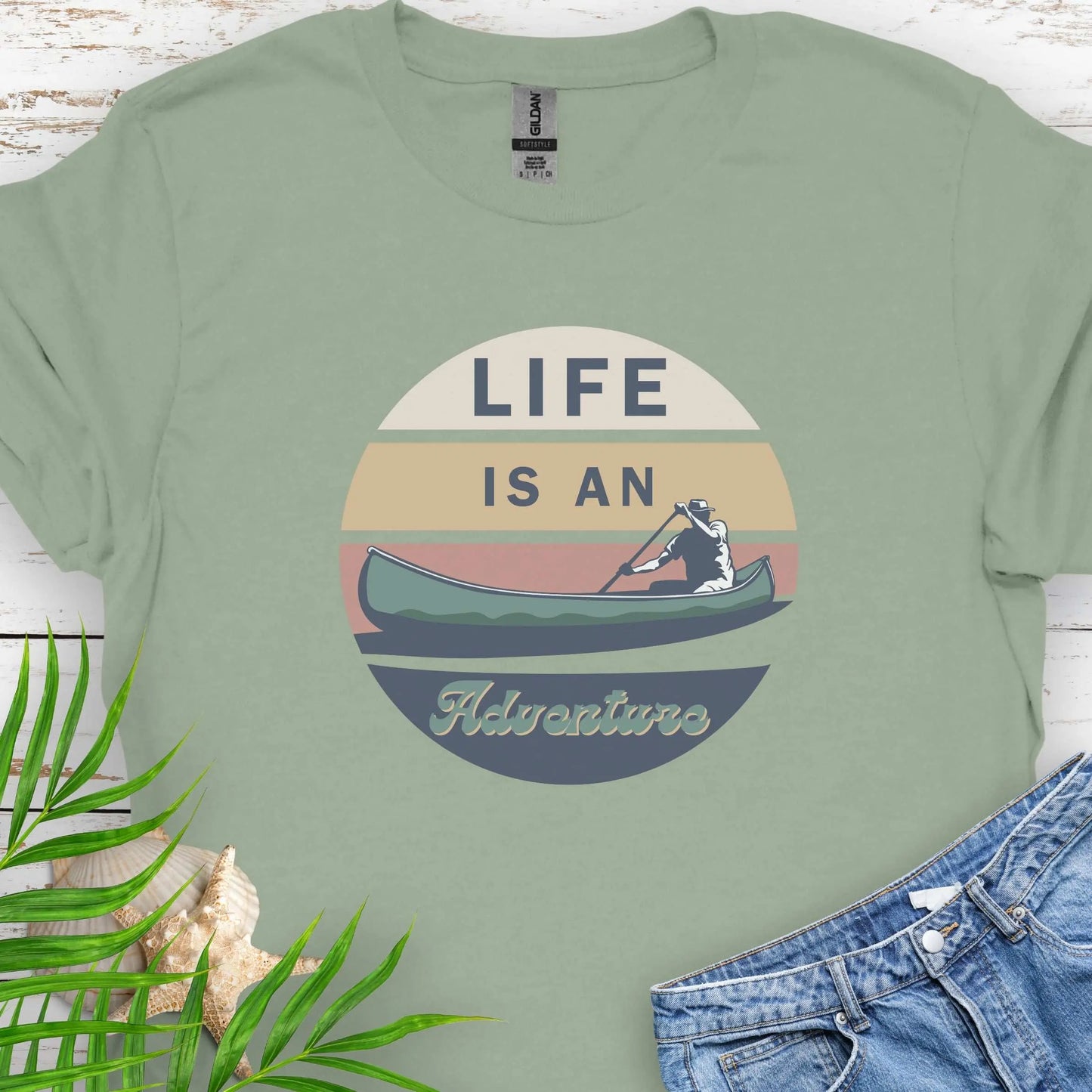 Sage green t-shirt featuring a retro sunset design with a man paddling a canoe and the phrase "Life is an Adventure" in bold, vintage-style font. Perfect for outdoor lovers, kayakers, and adventure seekers.