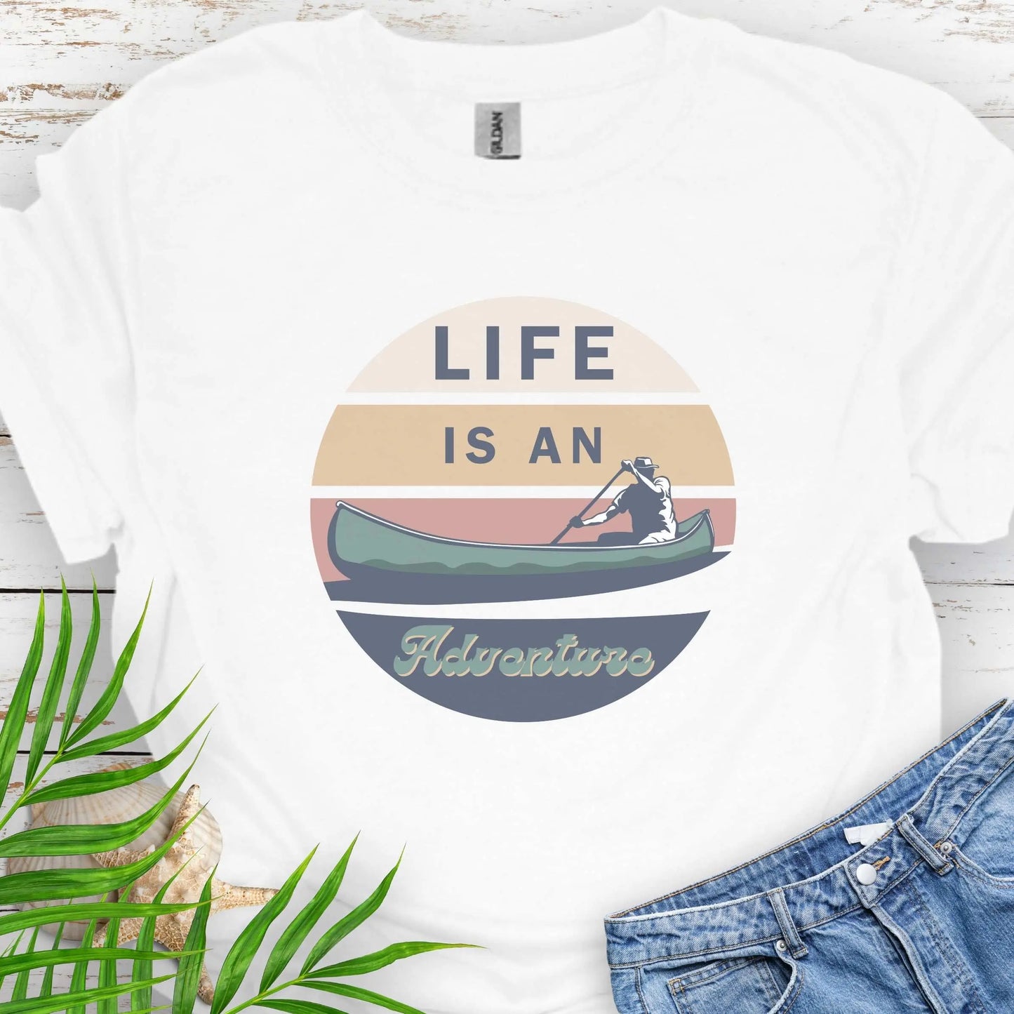 White t-shirt featuring a retro sunset design with a man paddling a canoe and the phrase "Life is an Adventure" in bold, vintage-style font. Perfect for outdoor lovers, kayakers, and adventure seekers.