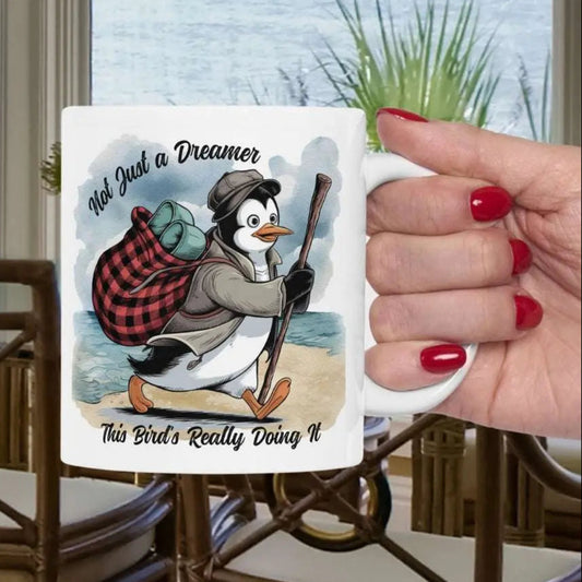 White 11 oz coffee mug featuring a fun beach design of a penguin running away on the sand with the text 'Not Just a Dreamer, This Bird’s Really Doing It.' A playful and unique gift for beach lovers and dreamers.