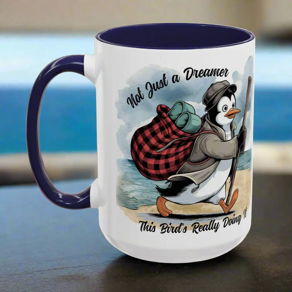 White 15 oz coffee mug with a navy blue interior and handle featuring a fun beach design of a penguin running away on the sand with the text 'Not Just a Dreamer, This Bird’s Really Doing It.' A playful and unique gift for beach lovers and dreamers.