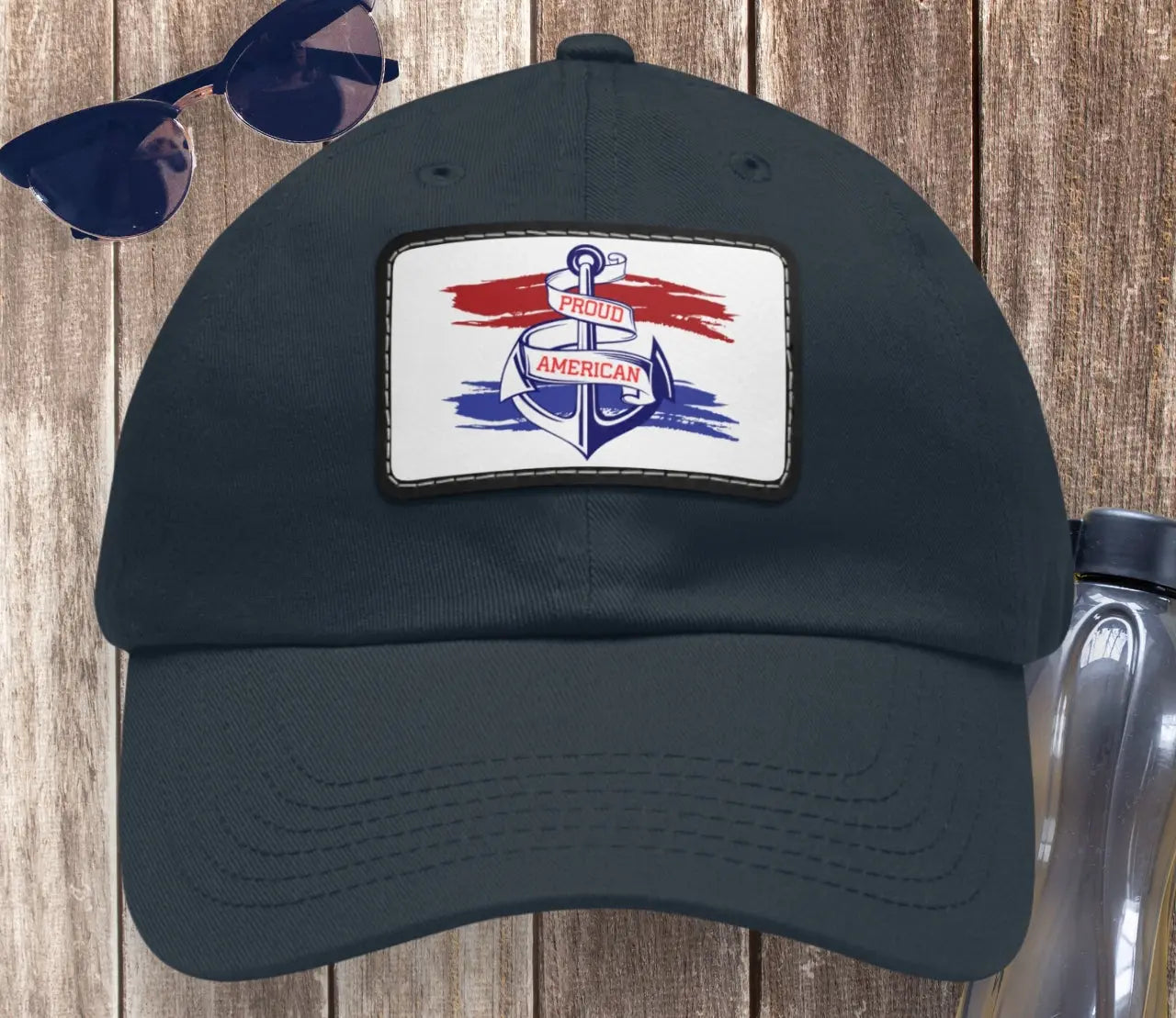 Flat view of Black hat with white patch red and blue behind a blue anchor with text Proud American of