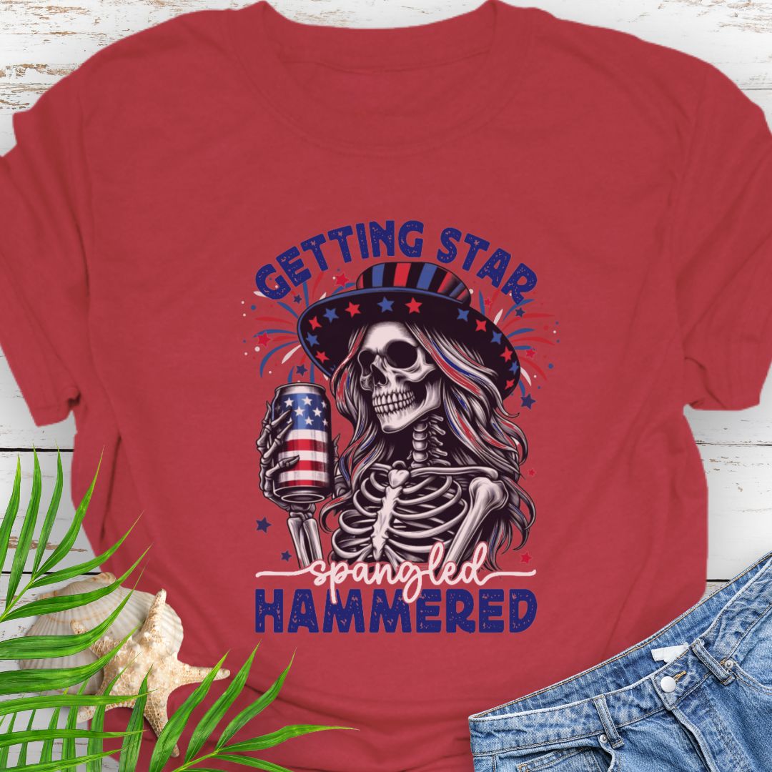 Cherry Red T-shirt graphic of a woman skeleton wearing a stars-and-stripes hat, holding an American flag beer can with the phrase “Getting Star Spangled Hammered.