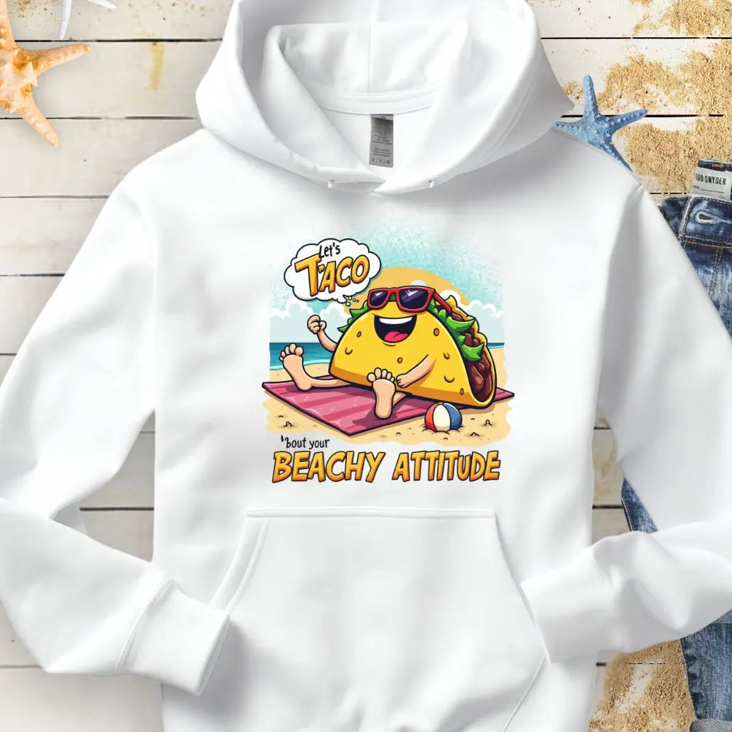 White hoodie with a taco wearing sunglasses laying on a beach towel on the beach with the text Let's Taco 'bout your Beachy Attitude.