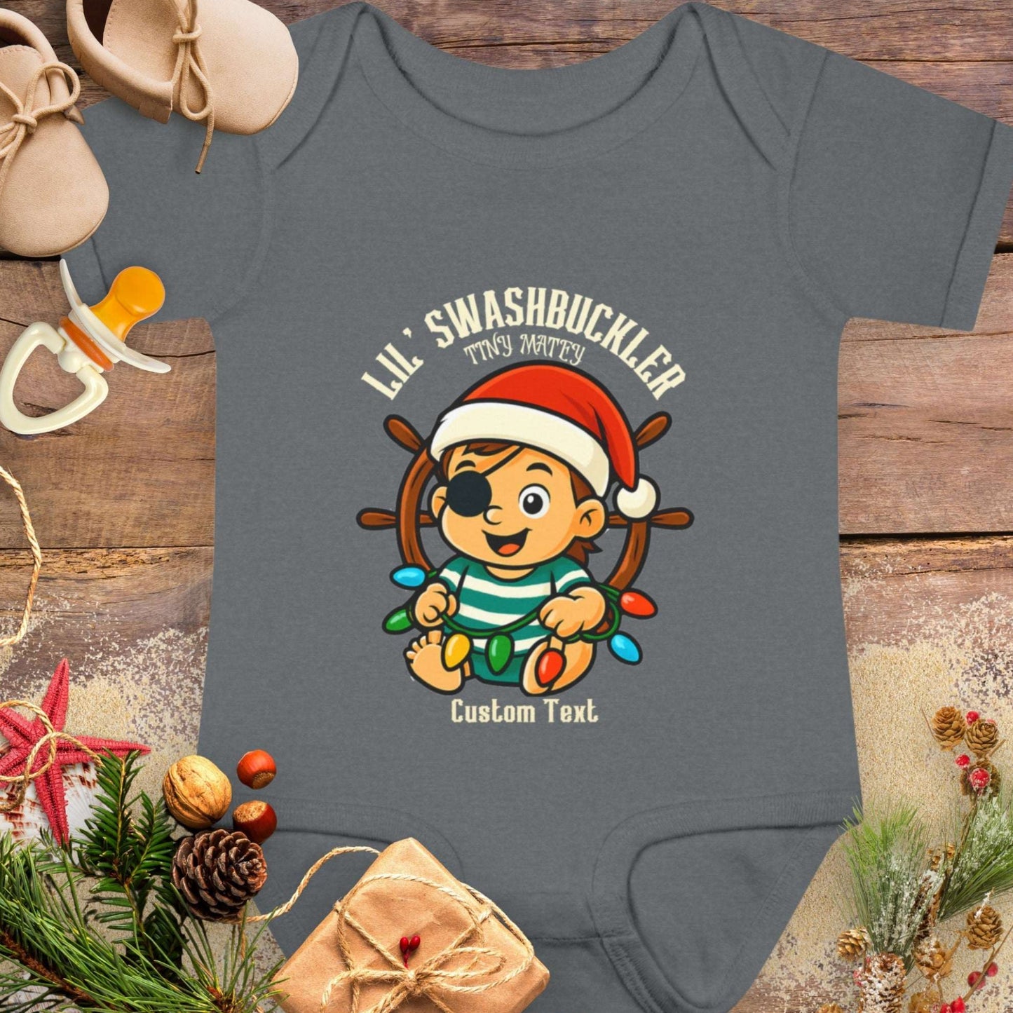 Gray baby onesie with Christmas-themed design featuring a cartoon baby pirate character and text 'Lil Swashbuckler Tiny Matey' that can be personalized.