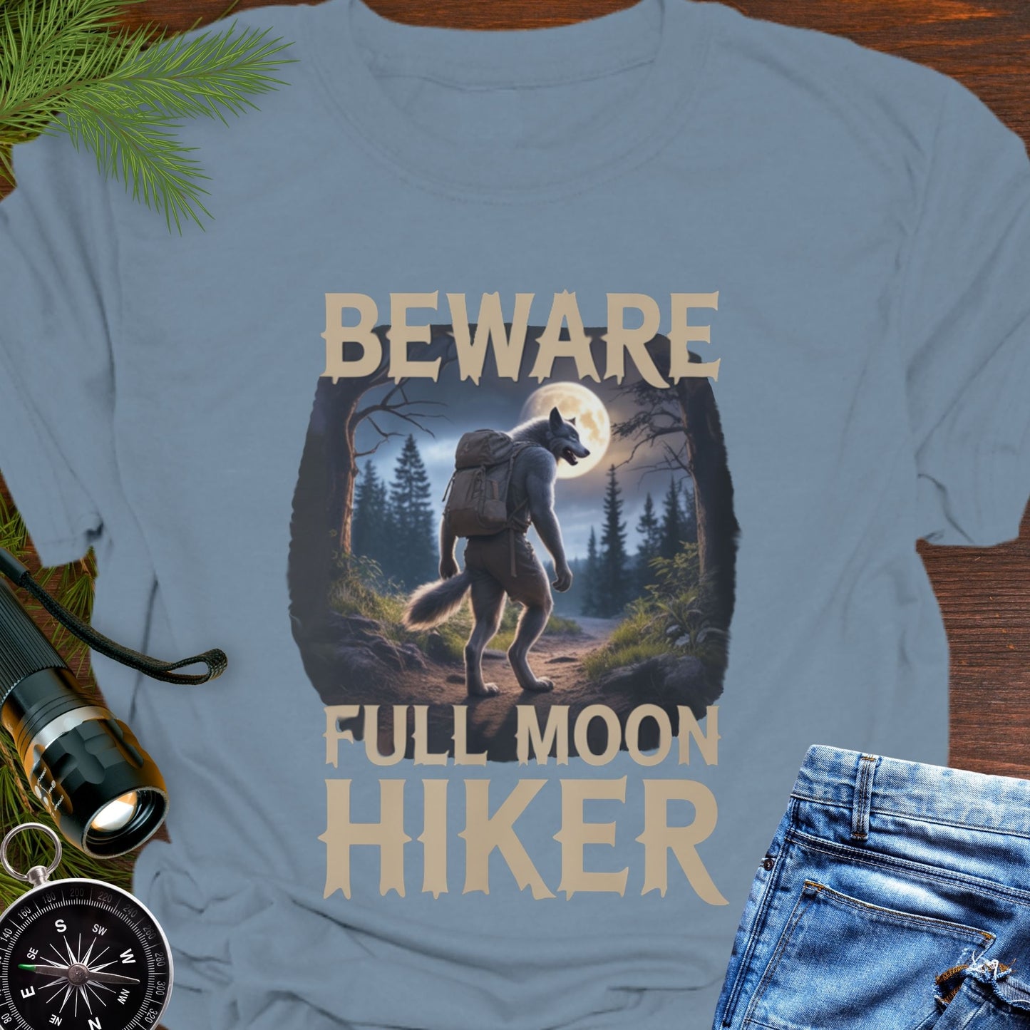 Light blue t-shirt with a graphic of a bear and text 'Beware Full Moon Hiker' on a wooden surface.