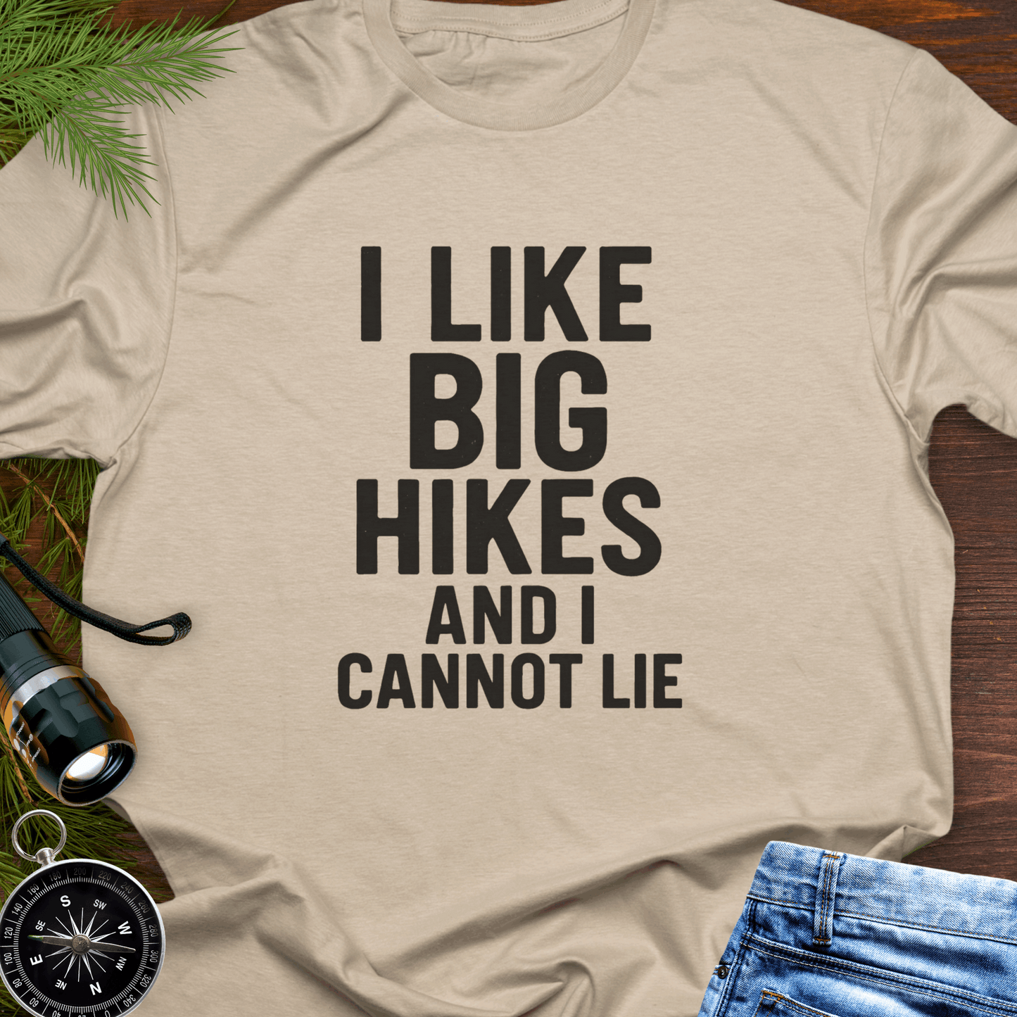 Beige t-shirt with 'I LIKE BIG HIKES AND I CANNOT LIE' text, surrounded by hiking accessories on a wooden surface.
