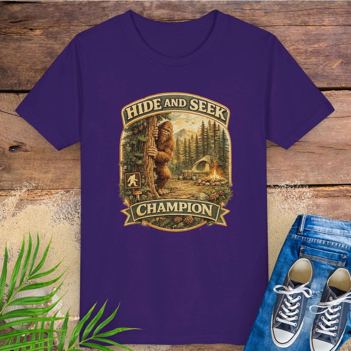 Purple t-shirt with 'Hide and Seek Champion' graphic with Bigfoot peeking around a tree in a campsite.