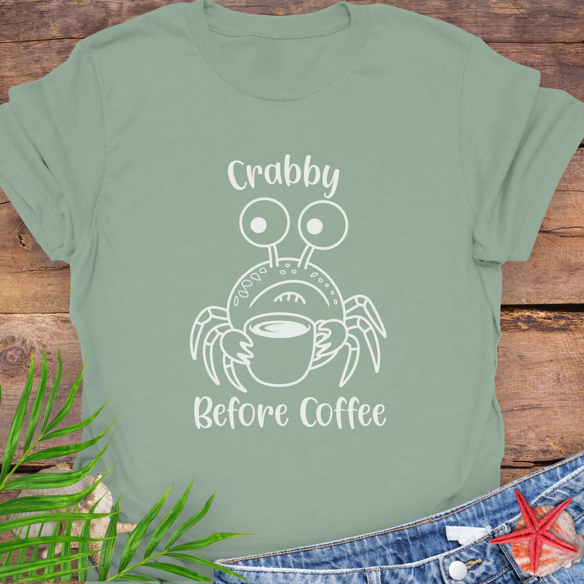 Green t-shirt with a crab graphic and text 'Crabby Before Coffee' on a wooden surface.