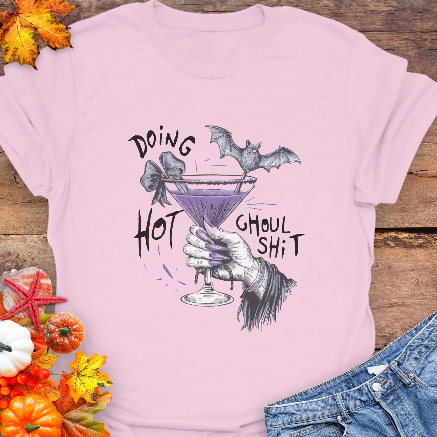 Pink t-shirt with Halloween-themed design featuring a purple cocktail and bat, on a wooden surface with autumn leaves and jeans.