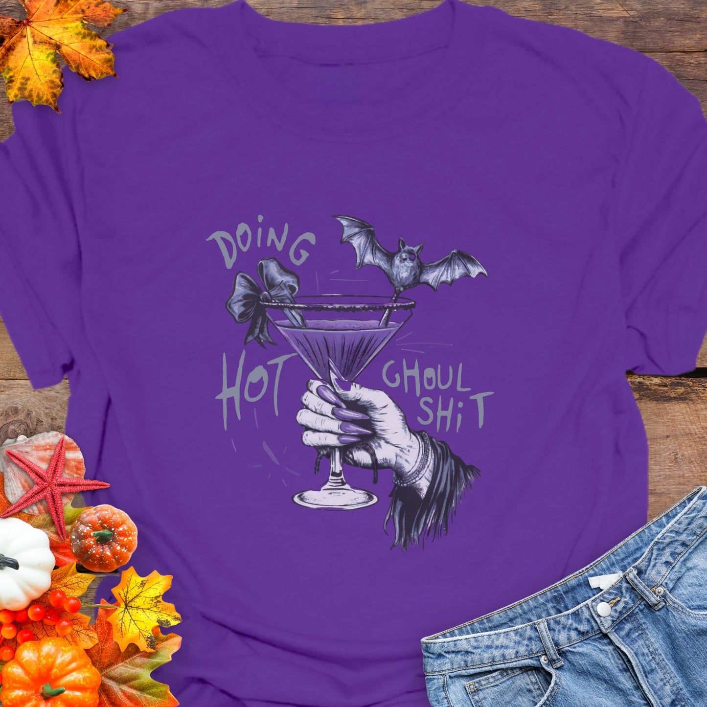 Purple t-shirt with a graphic design of a hand holding a martini glass with a bat on a wooden background.