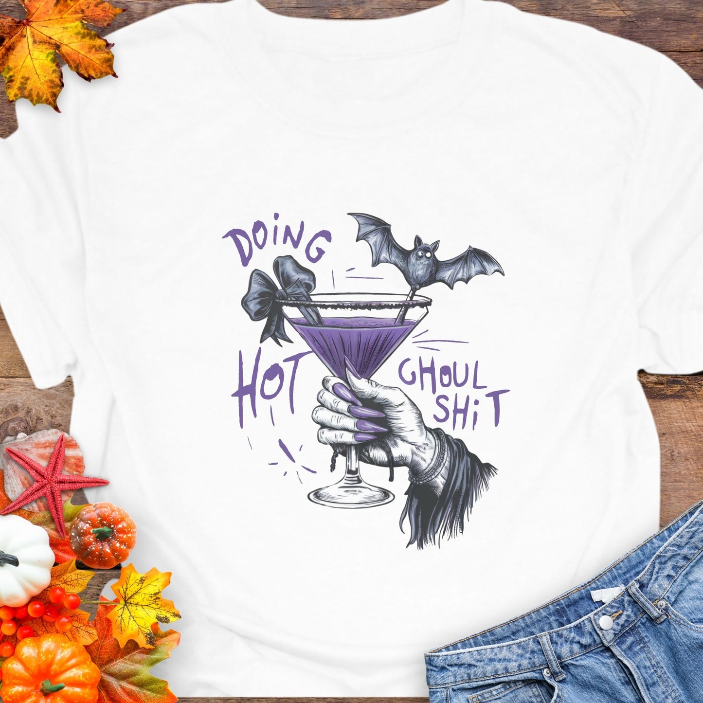 White t-shirt with Halloween-themed design featuring a bat, martini glass, and text on a wooden surface with autumn leaves and pumpkins.