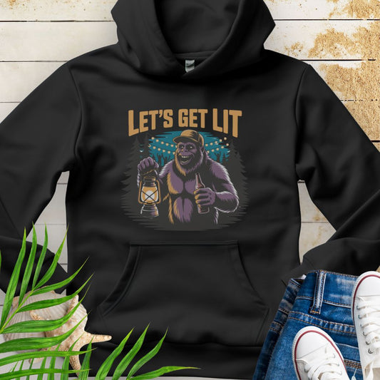 Black funny Sasquatch camping unisex hoodie with relaxed outdoor party vibe that says Let's Get Lit featuring a sasquatch wearing a ball cap holding a camping lantern and a bottle of beer with the forest in a forest camp scene.