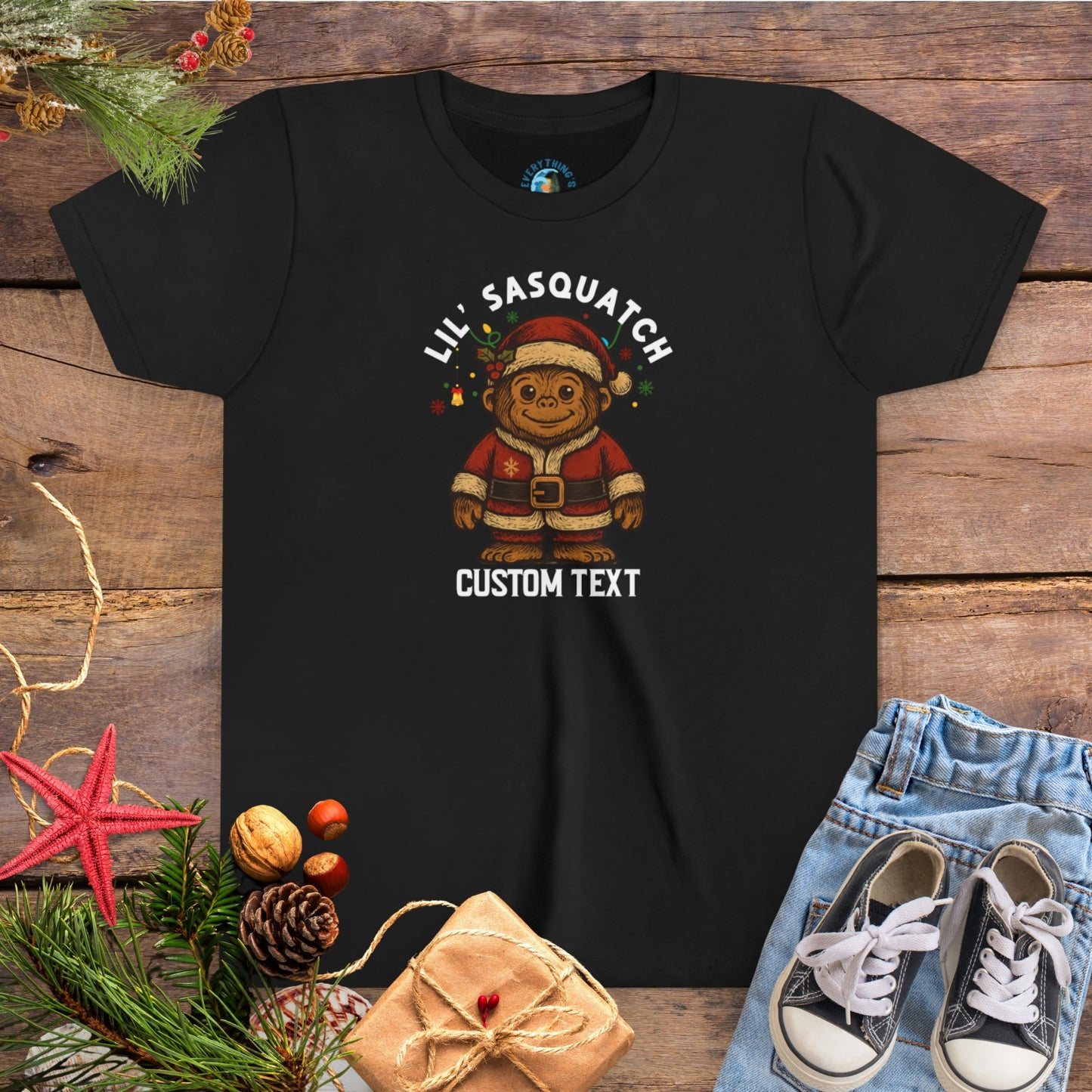 Black t-shirt with Christmas-themed graphic and 'Lil Sasquatch' text on a wooden surface with festive decorations.