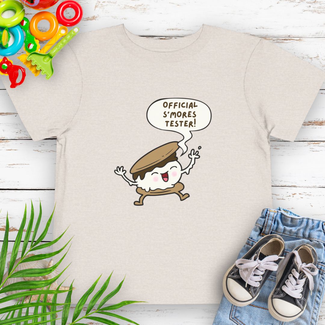 Heather Dust Toddler T-shirt with "Official S'mores Tester" text in speech bubble over a S'more character – perfect for camping and summer fun.