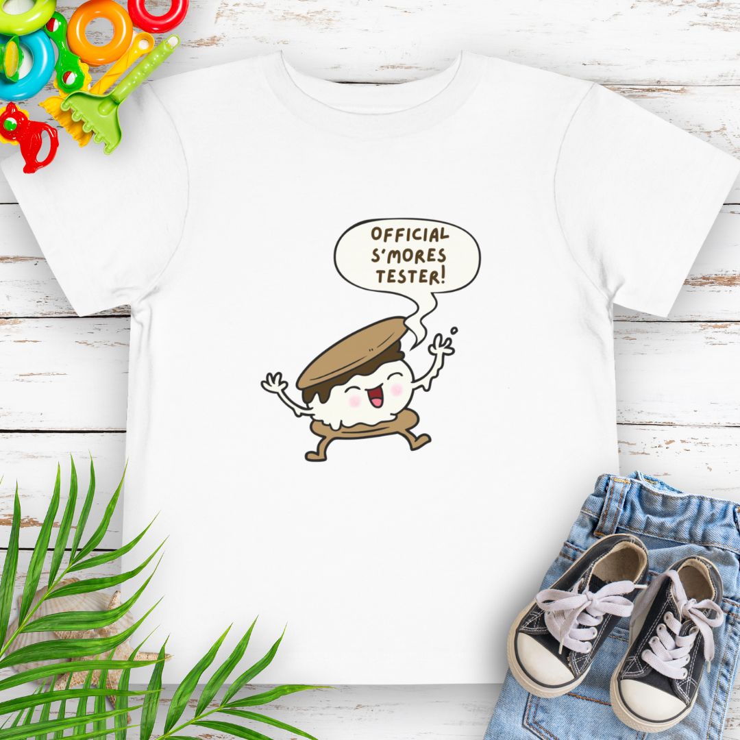 White Toddler T-shirt with "Official S'mores Tester" text in speech bubble over a S'more character – perfect for camping and summer fun.