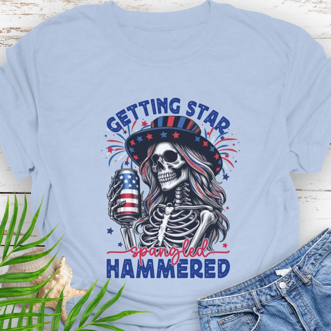 Light Blue T-shirt graphic of a woman skeleton wearing a stars-and-stripes hat, holding an American flag beer can with the phrase “Getting Star Spangled Hammered.