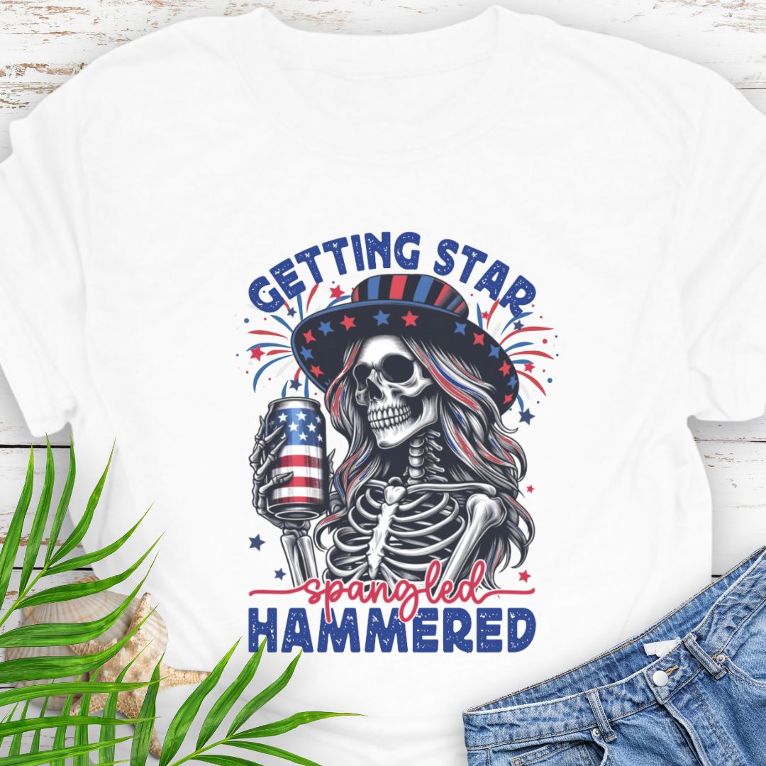 White T-shirt graphic of a woman skeleton wearing a stars-and-stripes hat, holding an American flag beer can with the phrase “Getting Star Spangled Hammered.