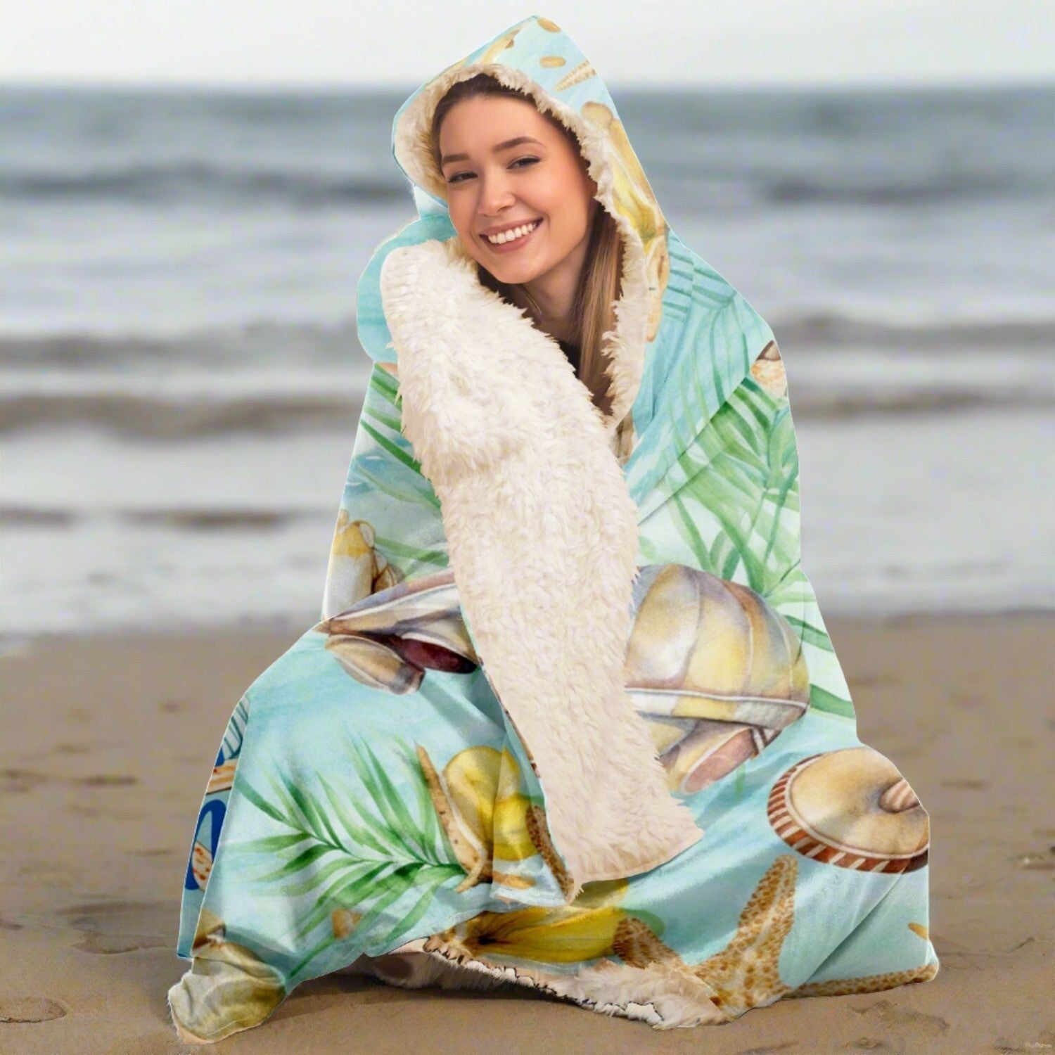 Person sitting on a beach wrapped up in a hooded blanket with a light aqua background with seashells and leaves with blue text that says Chill at the Beach.