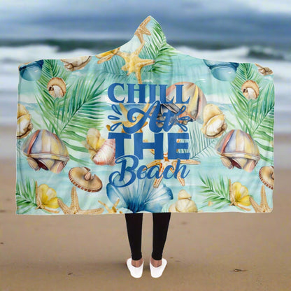 Person standing on a beach holding open a hooded blanket with a light aqua background with seashells and leaves with blue text that says Chill at the Beach.