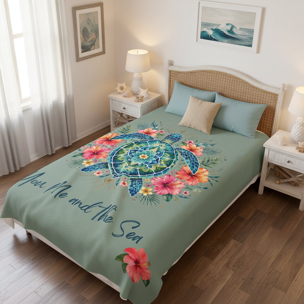 A sage green blanket featuring a colorful mandala-style sea turtle design surrounded by pink and orange hibiscus flowers, with the phrase 'You Me and the Sea' in dark blue script, shown on a bed in a bedroom.