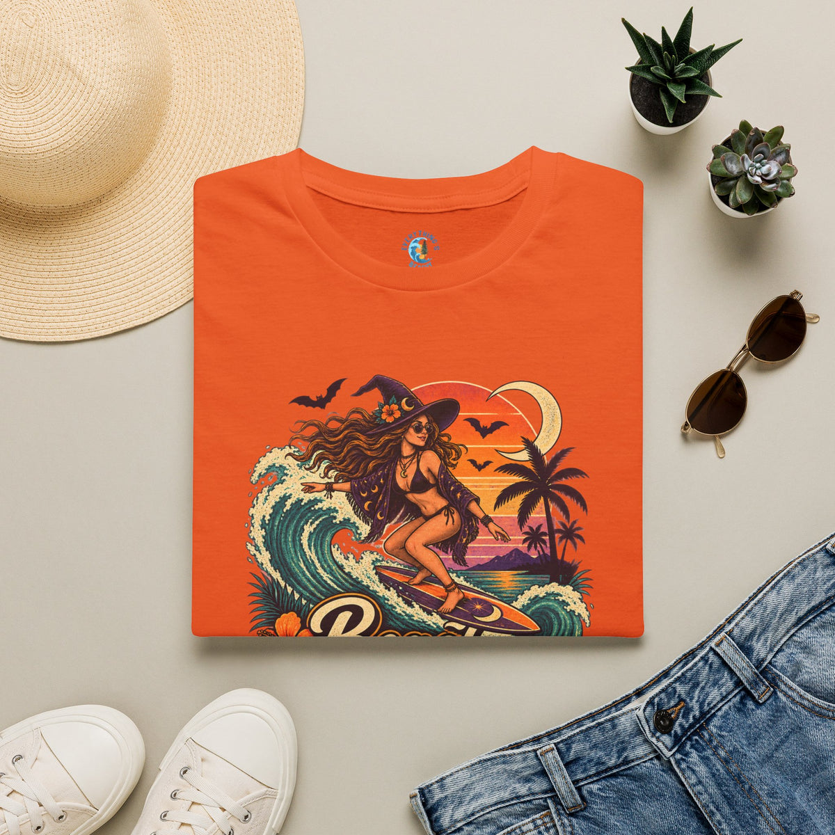 Folded Orange unisex t-shirt with “Beach Witch Energy” text, a witchy surfer girl, ocean wave, sunset, crescent moon, bats, palm trees, and tropical flowers.