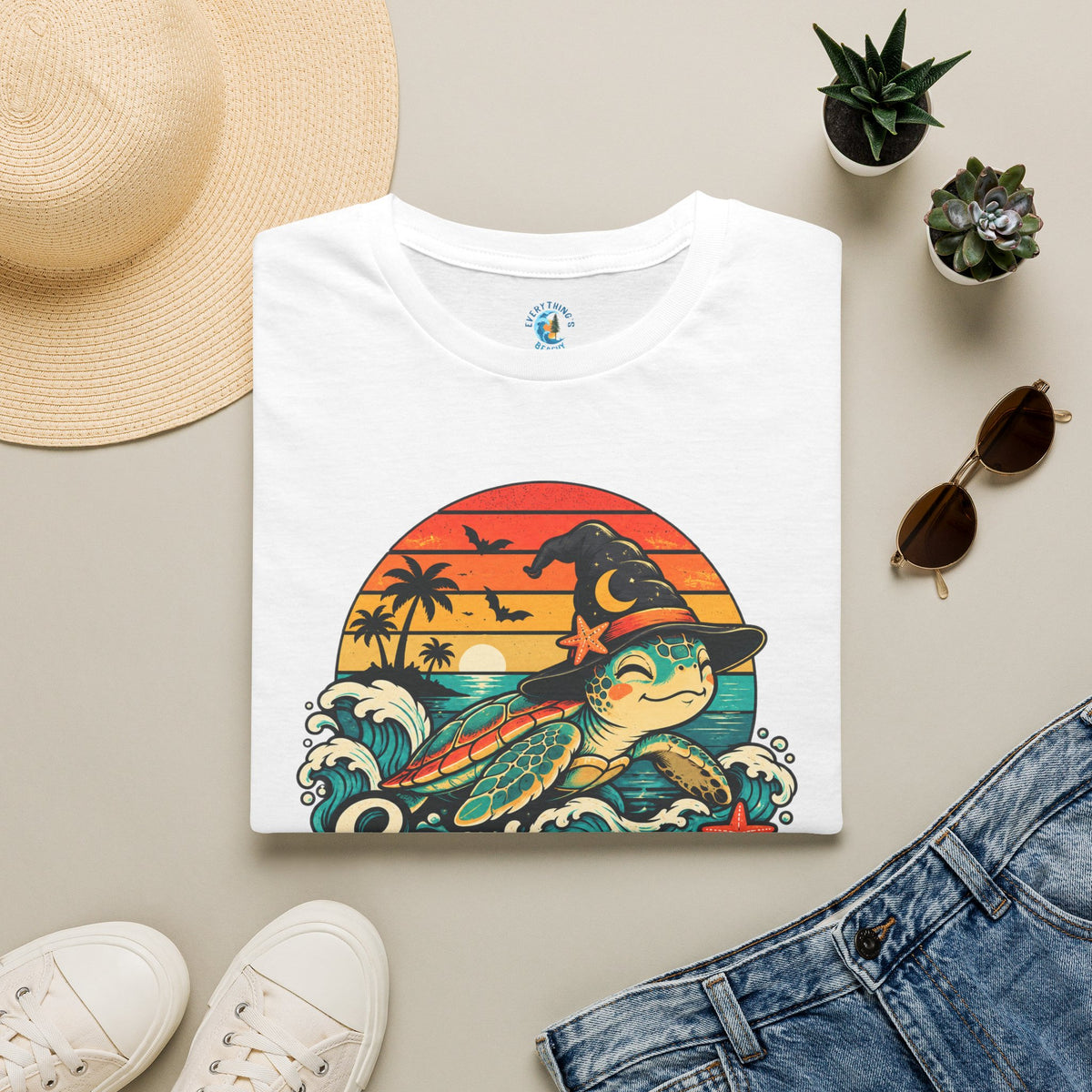 Folded White unisex t-shirt with “Seas the Magic” text, a sea turtle wearing a witch hat, ocean waves, sunset, palm trees, bats, and starfish.