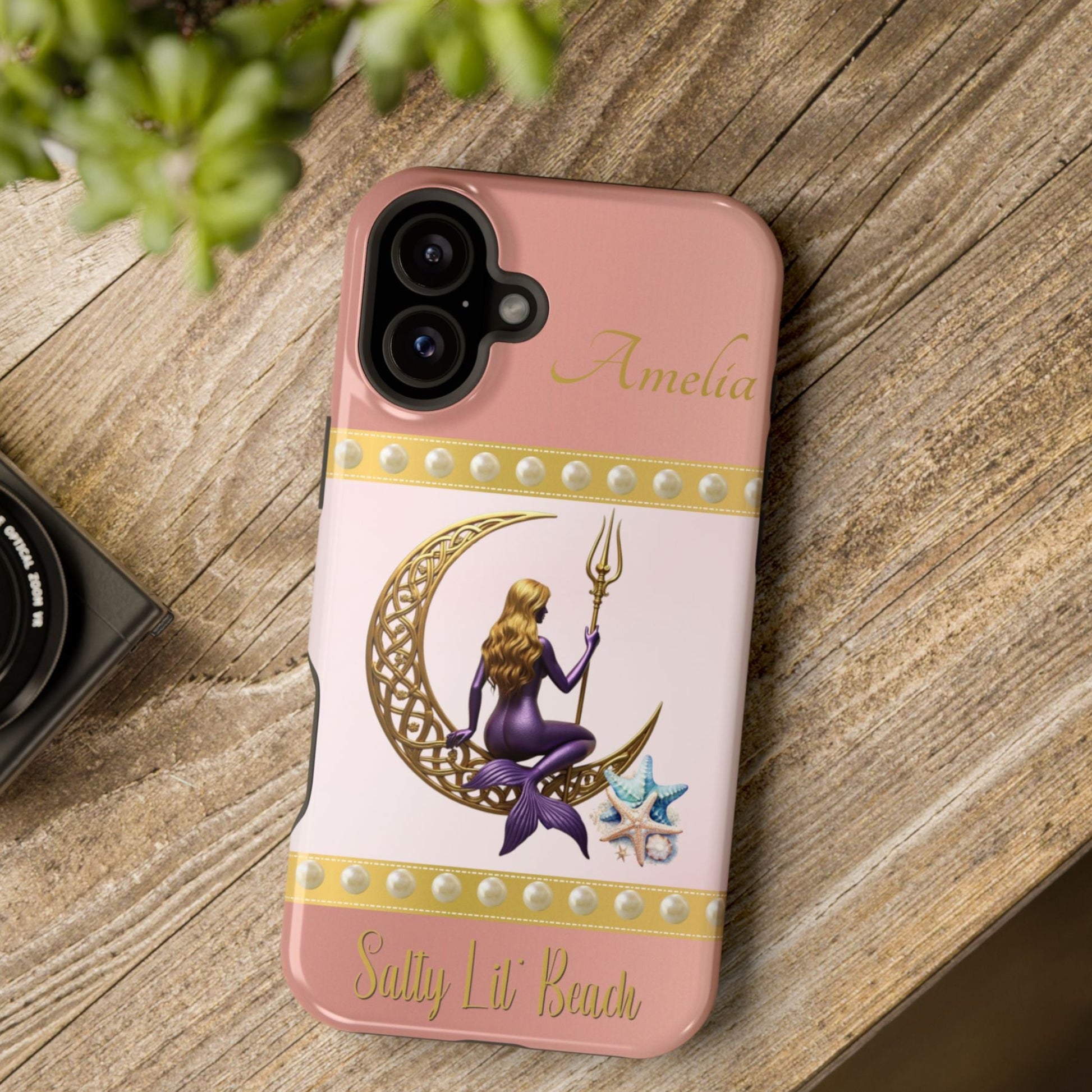 Phone case with a design of a mermaid and crescent moon on a pink background, placed on a wooden surface.