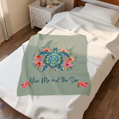 A sage green throw blanket draped over a bed, featuring a colorful mandala-style sea turtle design surrounded by pink and orange tropical flowers, with the text 'You Me and the Sea' in navy script below. The scene is set in a bedroom with wooden flooring and a wooden nightstand.