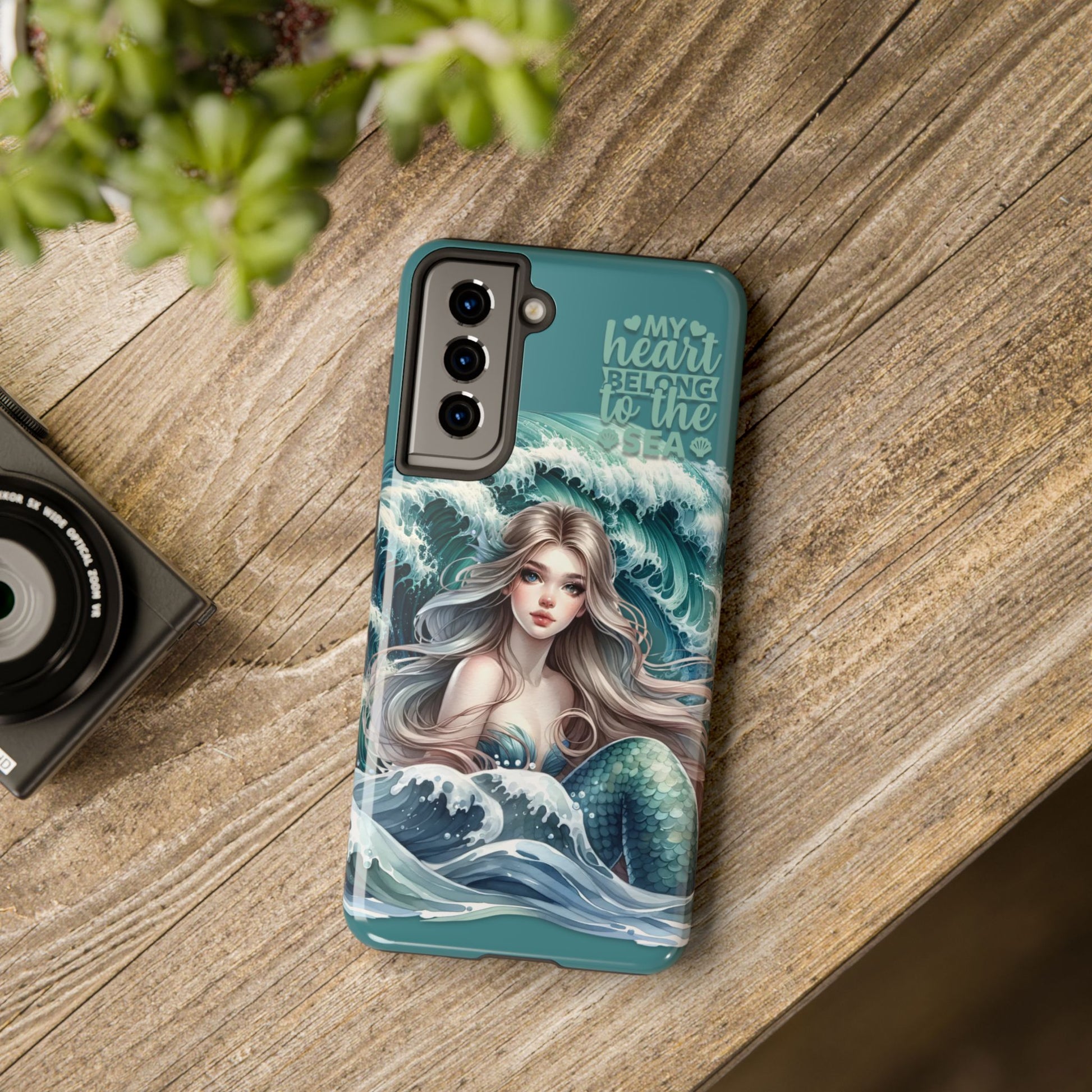 Phone Case with Flowy mermaid with ocean wave behind and text My Heart Belongs to the Sea laying on desk.