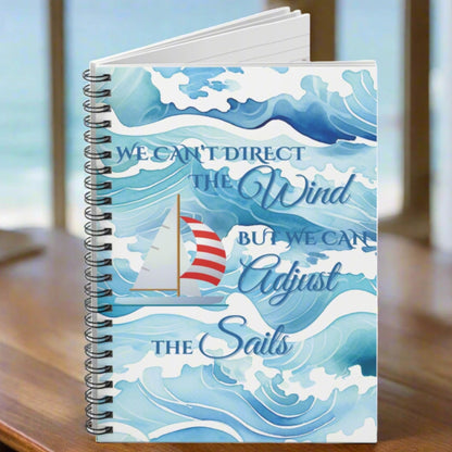 Notebook with blue and white ocean waves with a sailboat with red and white striped sails with the text We Can't Direct the Wind But we can Adjust the Sails